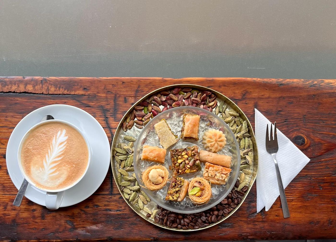 Cappuccino neben einem Messingteller mit Baklava, Pistazien, Kardamom und Kaffeebohnen auf der Holztheke am Fenster
