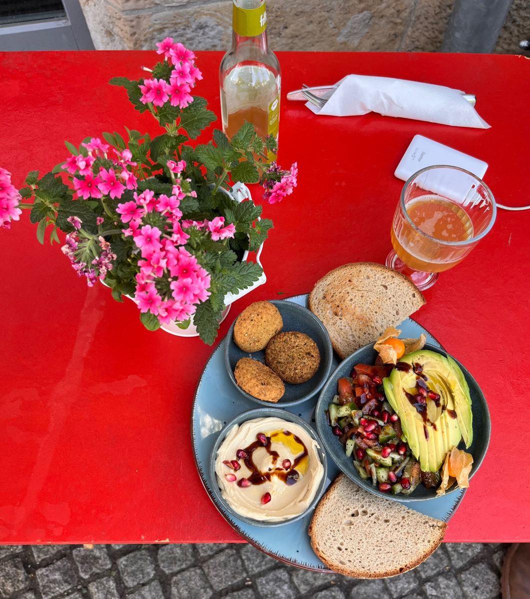 Roter Tisch draußen auf dem Kopfsteinpflaster mit Blumen, Limonade und einem Teller mit Falafel, Hummus und Salat