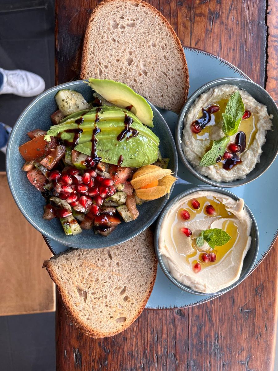 Teller mit Hummus, Baba Ghanoush, Salat mit Avocado und Granatapfel, dazu zwei Scheiben Brot auf altem Holz